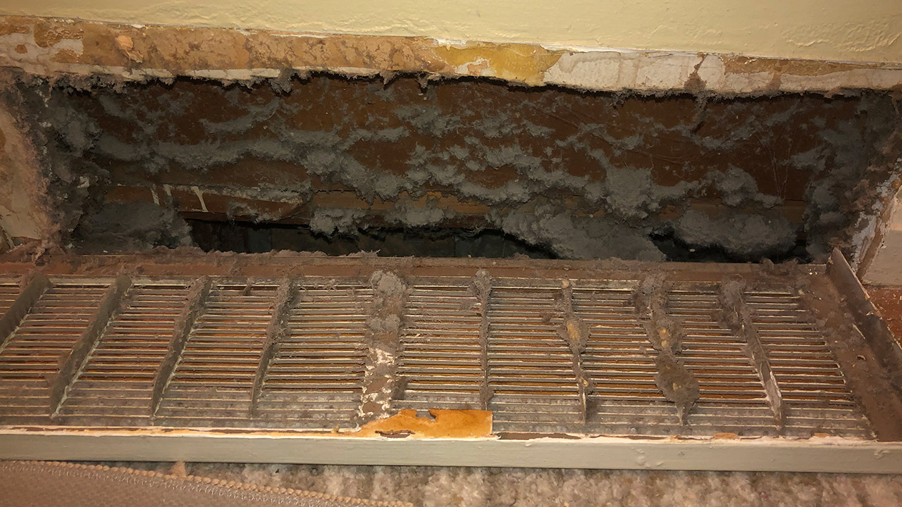 A dusty and dirty air vent filled with thick layers of dust, dirt, and debris surrounding the opening and grille.